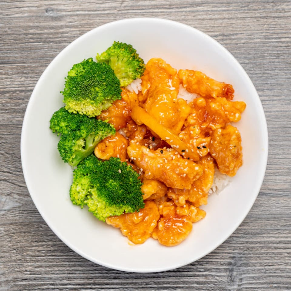 Orange chicken with sesame seeds, white rice, and broccoli florets in a white bowl.